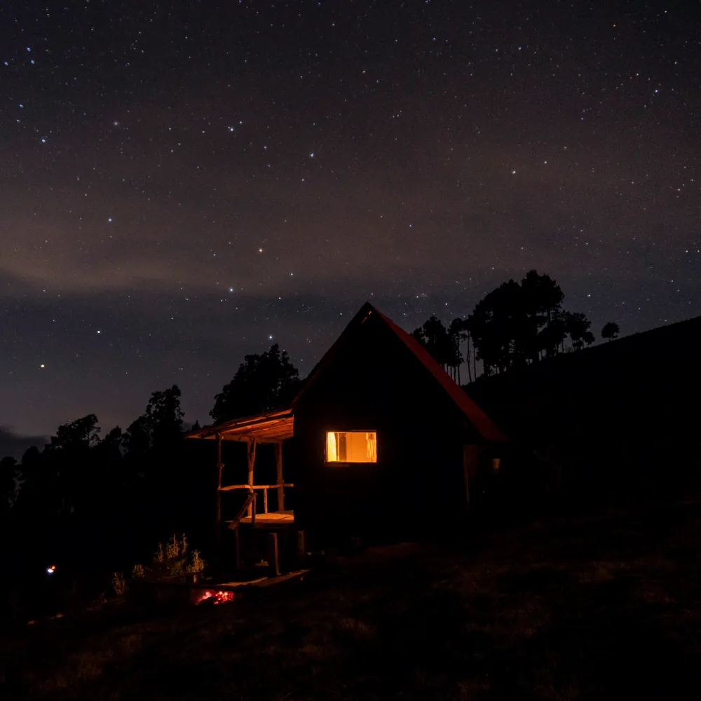 A cabin with one lit window on a dark hillside under the stars